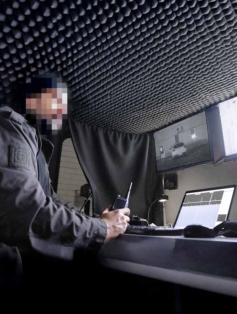 A private detective is sitting in a surveillance van, holding a walkie-talkie and watching the monitors.