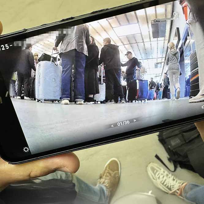 A smartphone shows a low-angle shot from a hidden airport camera: a long queue of travelers with wheeled suitcases under strip lighting, grey floor in focus. Gallery reads 01/36; a gear icon sits bottom-left. Offscreen shoes and a black backpack frame the scene. A private detective surveils passengers boarding the plane.